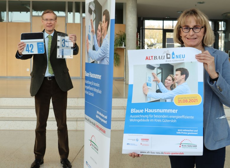 Landrat Sven-Georg Adenauer hält die Blauen Hausnummern in der Hand, die an die ausgezeichneten Häuser angebracht werden können. Projektleiterin Ursula Thering hält ein Werbeplakat in den Händen. 