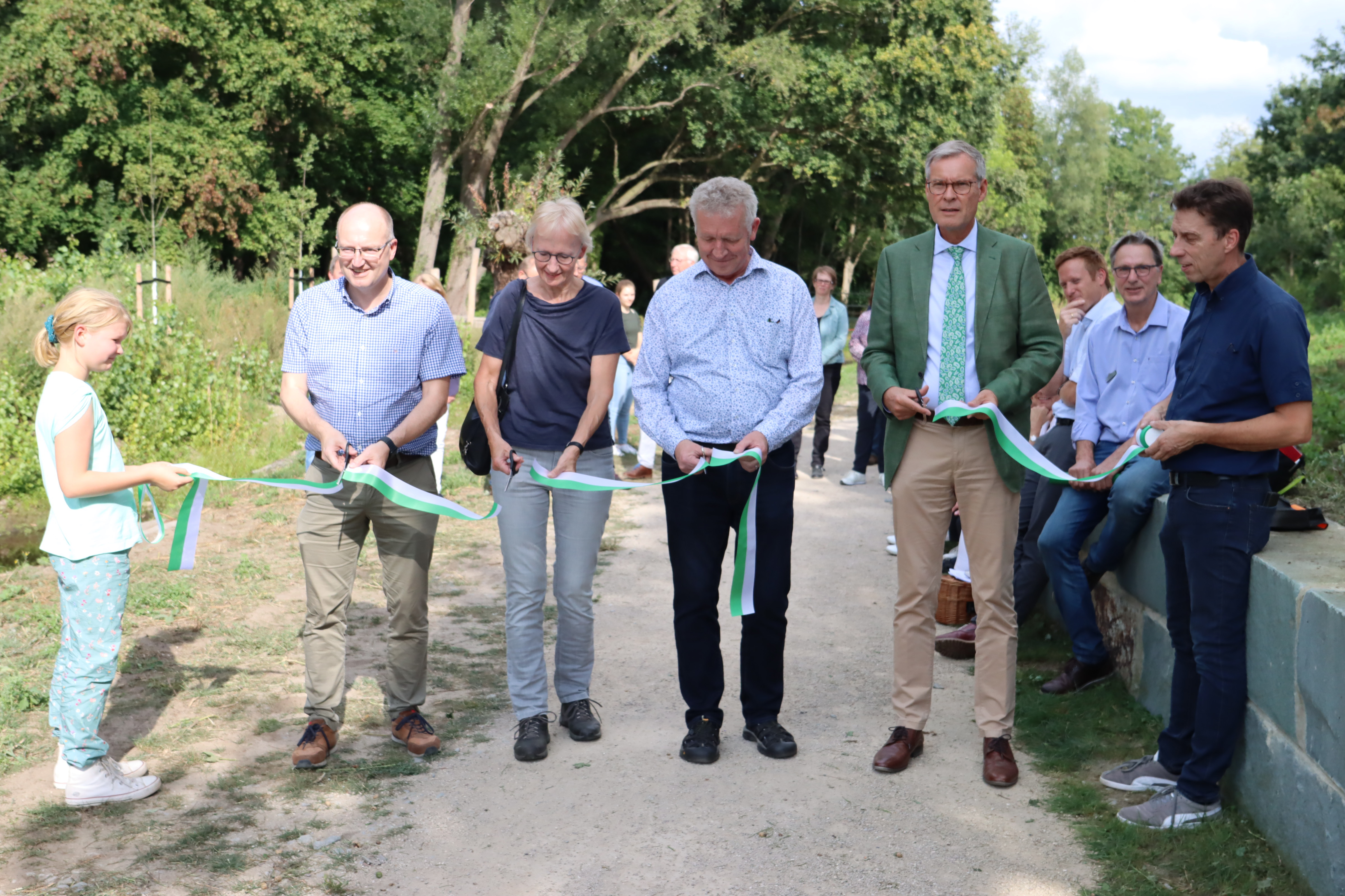(v.l.) Luzie hält das Band: Bürgermeister der Stadt Verl Michael Esken, Vorsitzende des Ausschusses für Klima- und Umwelt des Kreises Gütersloh Birgit Niemann-Hollatz (GRÜNE), Hans-Peter Jakobfeuerborn (Ratsmitglied Stadt Verl, CDU), Landrat Sven-Georg Adenauer und Bernd Meißner (Leitung Fachbereich Tiefbau, Stadt Verl) schneiden es durch. Band wird zur Eröffnung von fünf Personen durchgeschnitten. Ein Kind hält dies an einem Ende in den Händen.