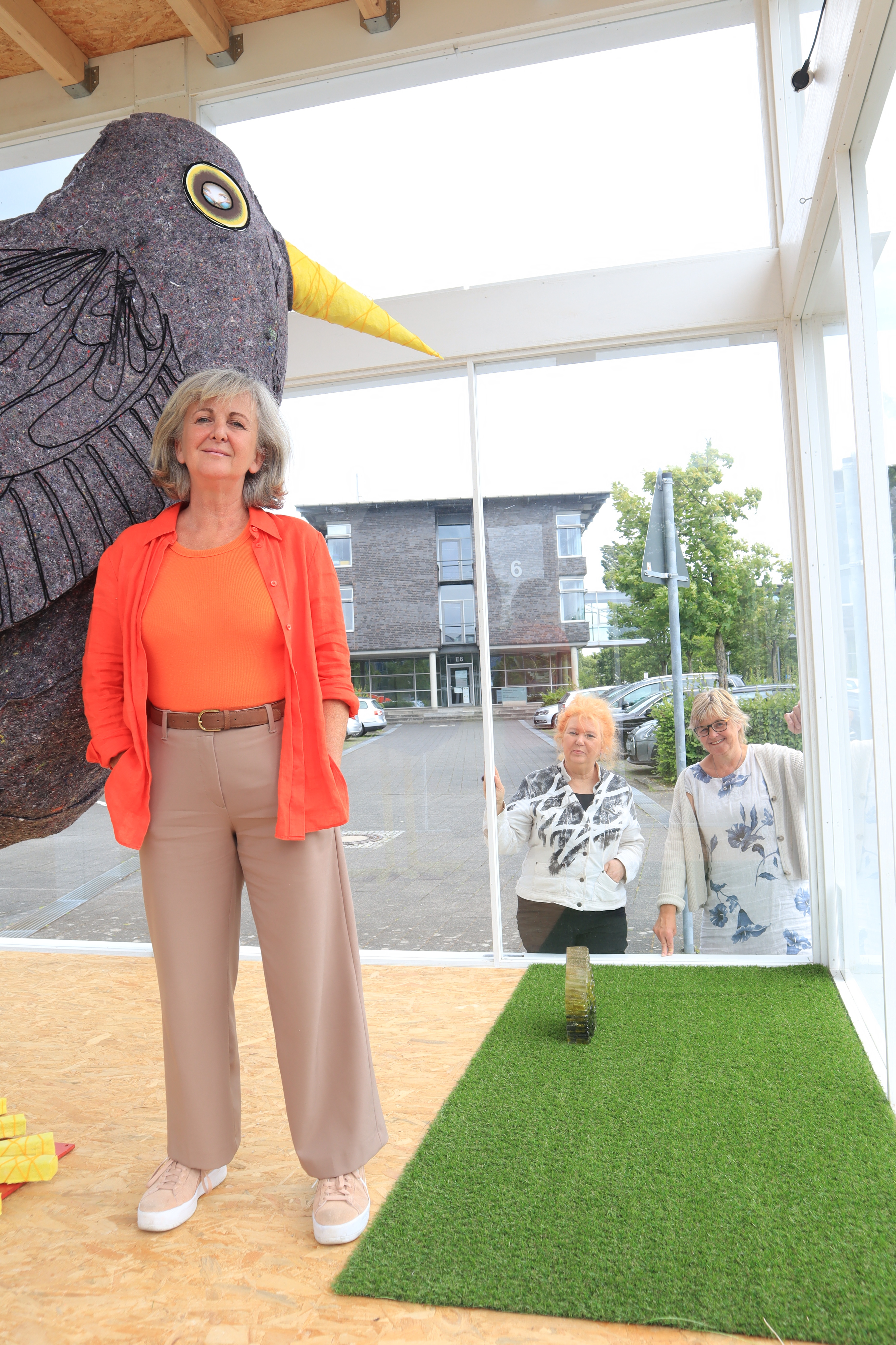 Die Juli-Ausstellung im Kunstpavillon ist eröffnet: (v.l.) Künstlerin Cristina Zanotti aus Schloß Holte-Stukenbrock mit Beate Freier-Bongaertz (Kunstverein Kreis Gütersloh) und Beate Behlert (Referat Presse, Kultur und Archiv des Kreises Gütersloh). In einem Glaspavillon ist ein großer, schwarzer Vogel zu sehen. Daneben steht die Künstlerin. Im Hintergrund sind zwei weitere Personen zu sehen.