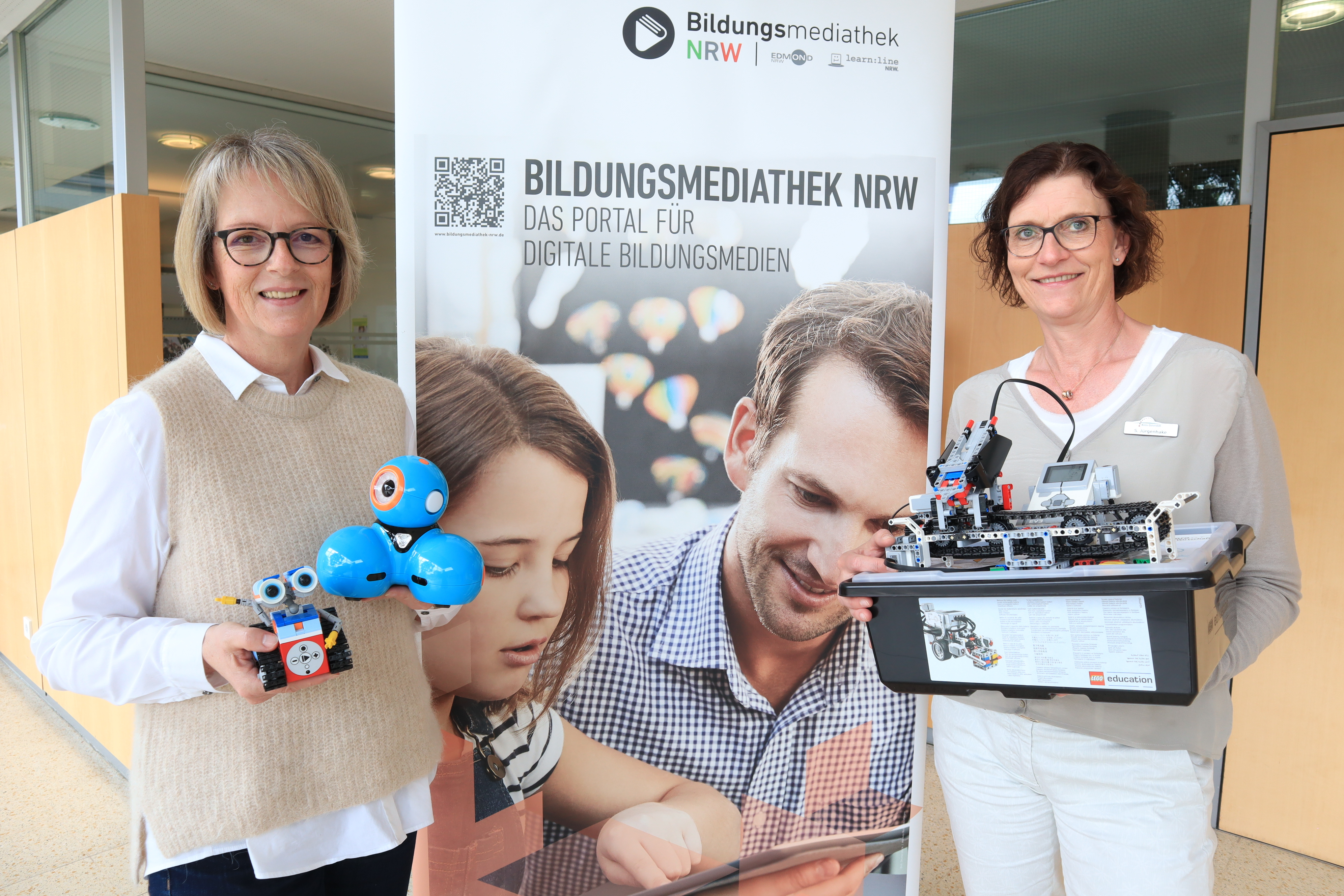 Beim Medienzentrum können kleine Roboter und Bausätze zum Programmieren für Kinder und Jugendliche ausgeliehen werden: Petra Poggenklas (links) hält den kleinen fahrbaren Roboter ‚Dash‘ in den Händen, Sandra Jürgenhake einen Bausatz für einen Roboter inklusive programmierbaren Computer. Zwei Personen, die vor einem Banner stehen. In den Händen halten sie einen kleinen Roboter und einen Bausatz in einem Karton.