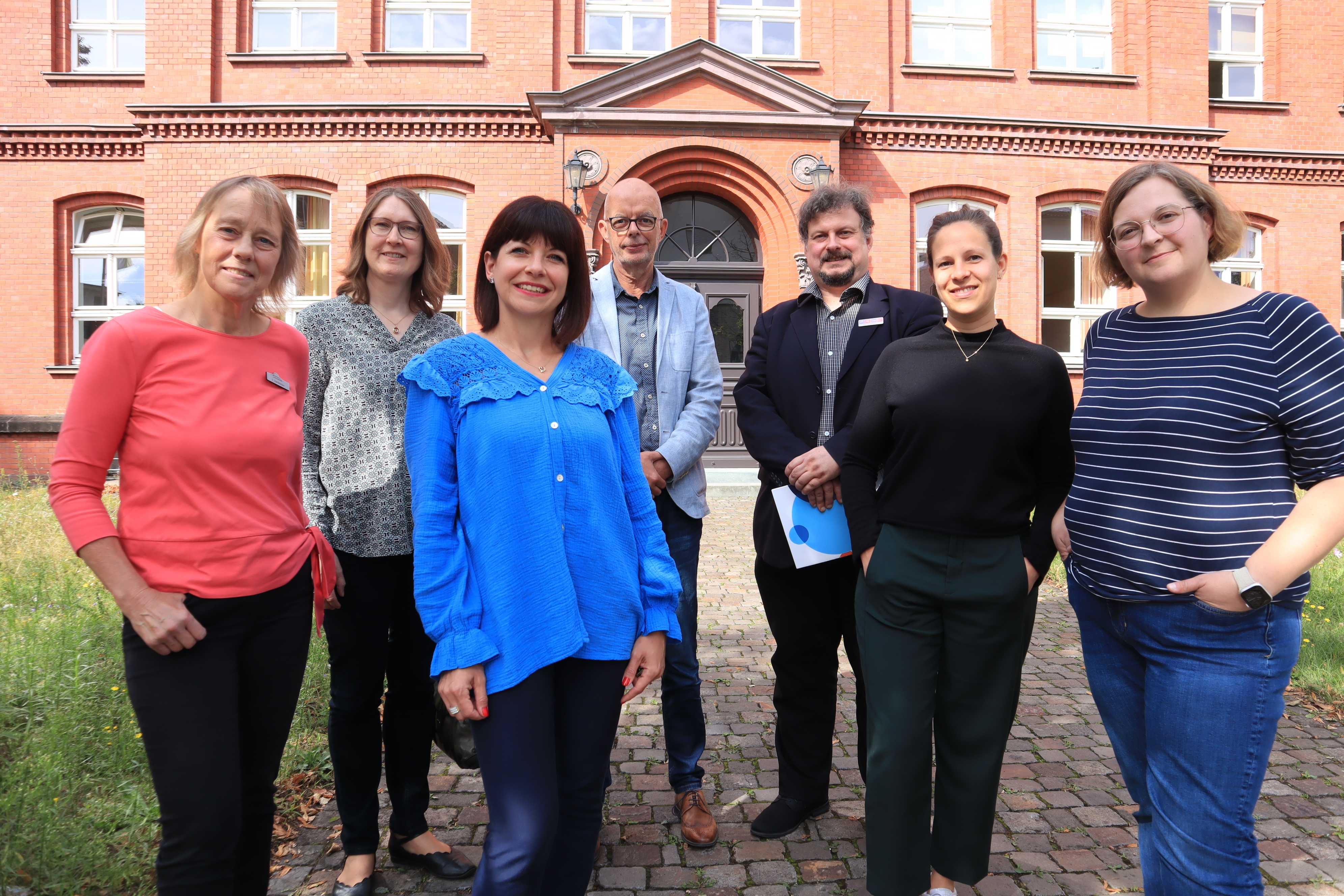 Kooperationspartner der Sprachcamps: (v.l.) Erika Dahlkötter (Kreis Gütersloh), Kreisdirektorin Susanne Koch, Andjelka Krstic und Dr. Elmar Schnücker (beide VHS Gütersloh), Axel Hemminghaus (VHS Reckenberg-Ems), Jelena Jaissle (VHS Verl, Harsewinkel, Schloß Holte-Stukenbrock) und Anna Behring (INTAL Bildung und Beruf gGmbH). Sieben Erwachsene, die vor der Volkshochschule Gütersloh stehen.