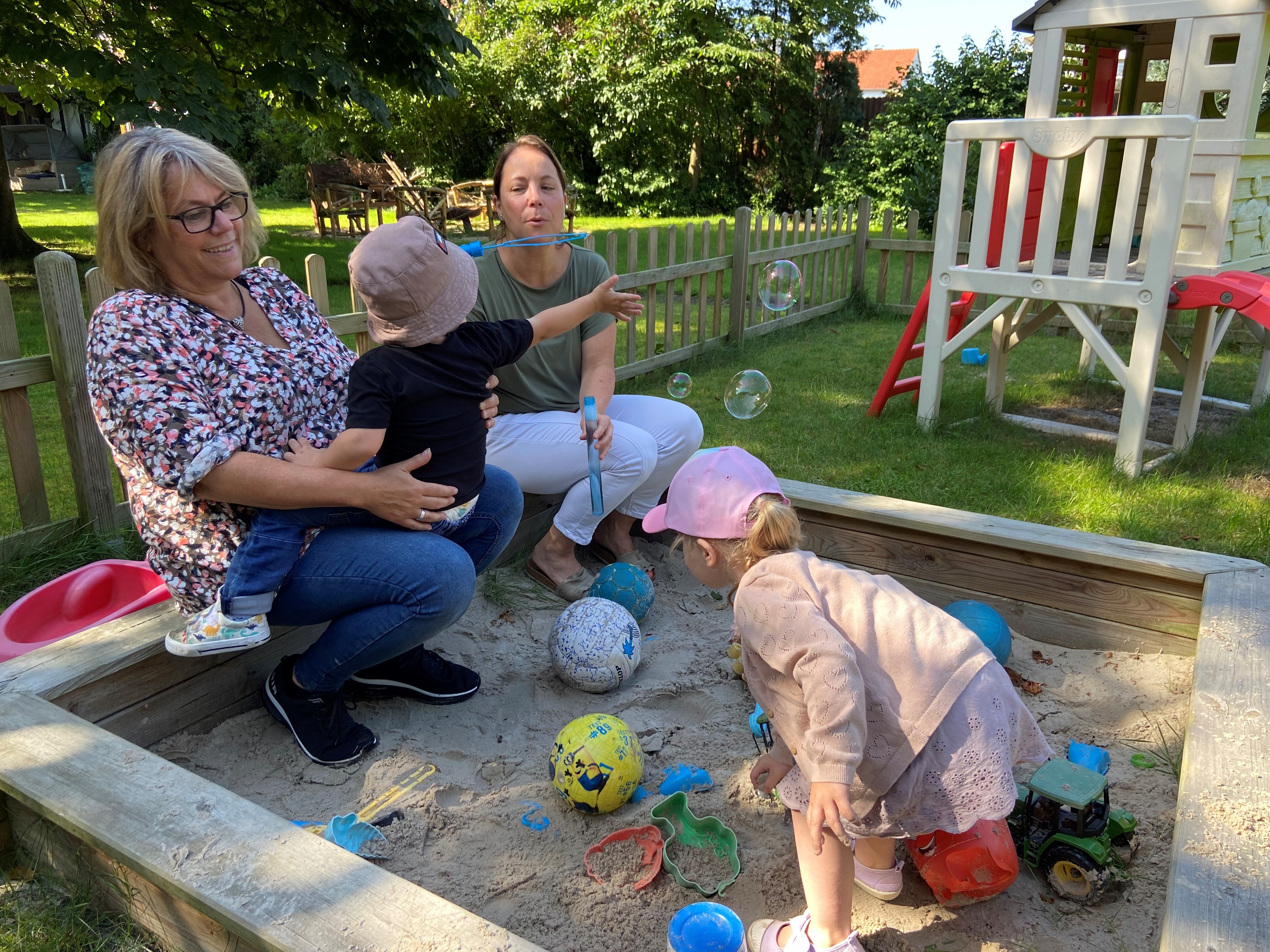 Lia und Louis machen mit ihrer Kindertagespflegeperson Heike Schmidt (links) und Kerstin Walter (Vermittlung für Kindertagespflege Versmold) Seifenblasen im Sandkasten. Zwei Erwachsene mit zwei Kindern im Sandkasten. Ein Kind sitzt auf dem Schoß, das andere spielt im Sand.