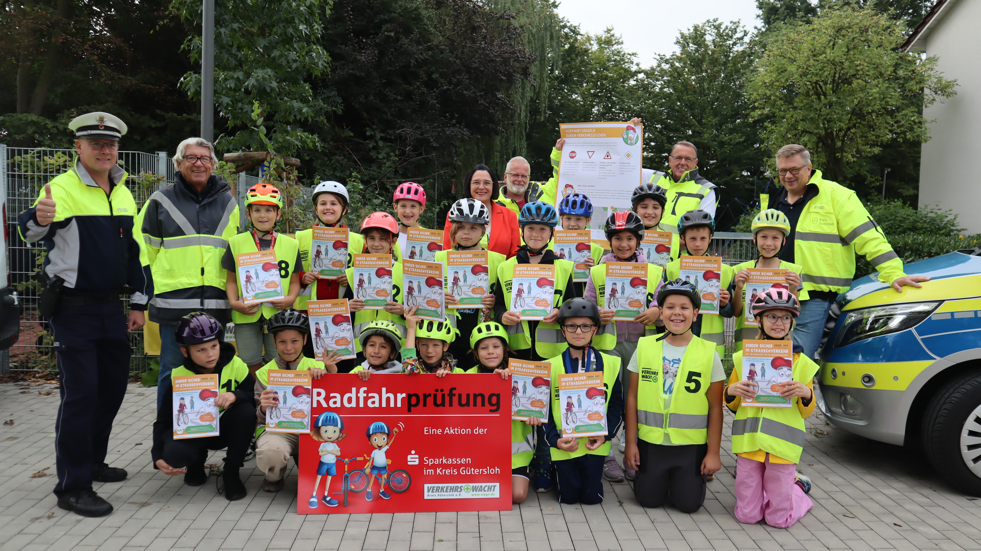 v.l.: Alexander Hüske (Polizeihauptkommissar Kreispolizei Gütersloh), Jür-gen Peta (Verkehrswacht), Regina Schneider (Marketing und Vertrieb K&L Verlag), Joachim Rothaus (Verkehrswacht), Bruno Wiemann (1. Vorsitzen-der Verkehrswacht Kreis Gütersloh e.V.) und Bernhard Riepe, (Leiter der Abteilung Straßenverkehr Kreis Gütersloh) mit den Kindern der Igel-Klasse. Die Verantwortlichen stehen mit den Schülerinnen und Schülern in gelben Warnwesten zusammen. Sie halten das Arbeitsheft hoch.