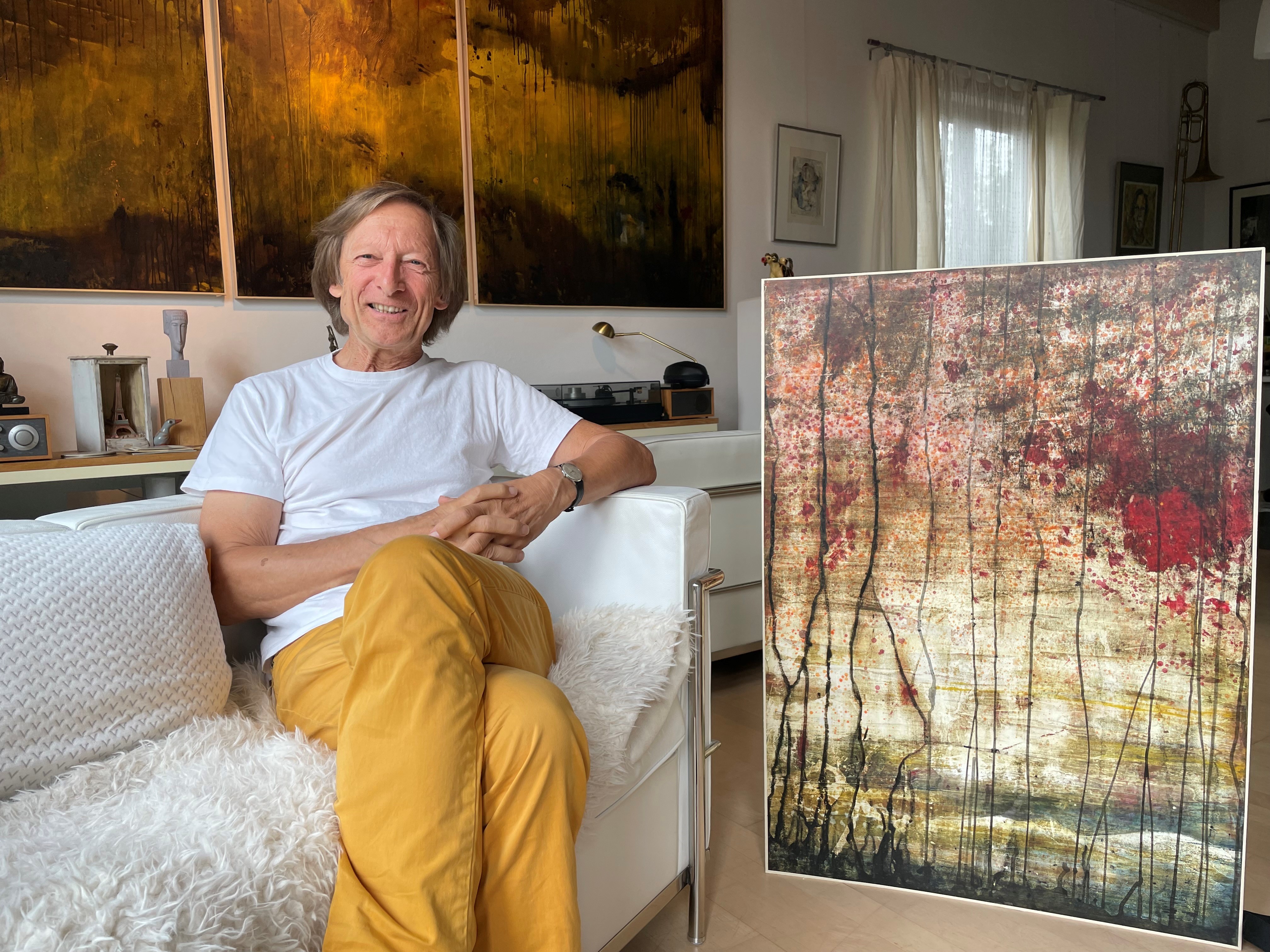 Künstler Karl-Heinz Lünstroth sitzend auf dem Sofa. Im Hintergrund ist seine Kunst zu sehen.