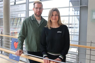 Ein Mann und eine Frau stehen im Kreishaus Gütersloh vor den Glasfronten und blicken in die Kamera. 