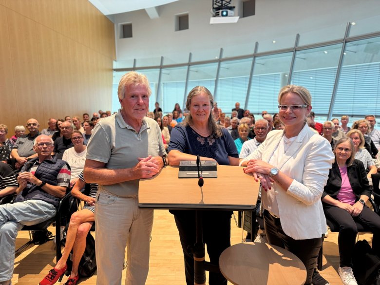 Drei Personen stehen am Pult, hinter ihnen sitzen die Ausstellerinnen und Aussteller.