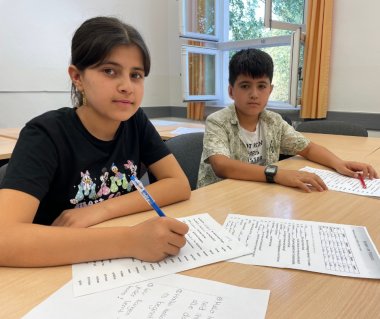 Zwei Kinder sitzen an Tischen und haben Zettel mit Aufgaben vor sich.