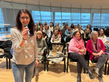 Eine Person steht vor einem Publikum, das im Hintergrund auf Stühlen sitzt. Die Referentin hält ein Mikrofon in der Hand. 