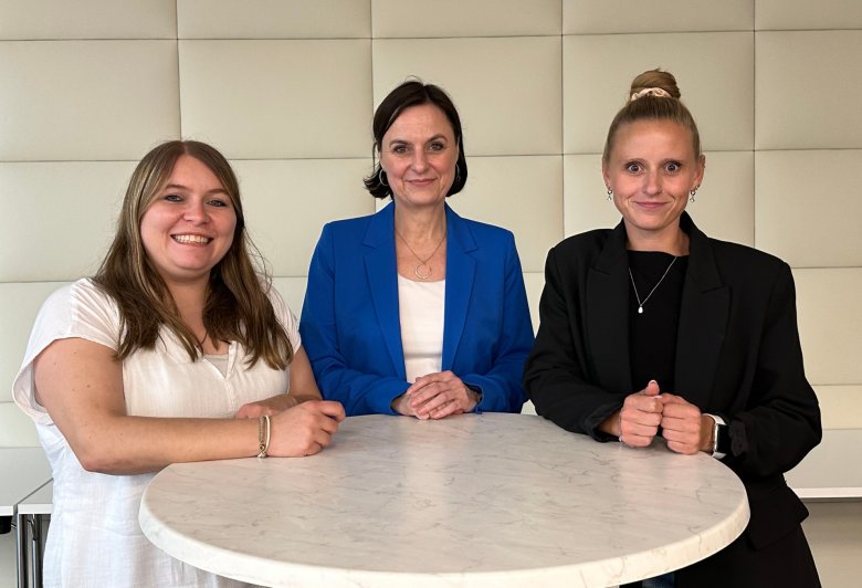 Drei Frauen stehen an einem Stehtisch nebeneinander und blicken in die Kamera.