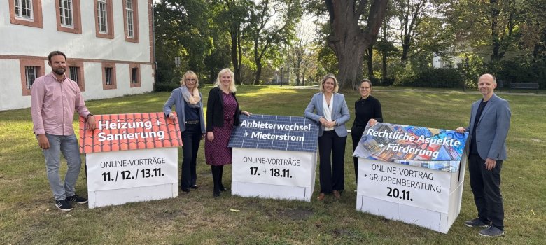 Personen stehen im Grünen, neben Papphäusern auf denen Daten und Titel der Vorträge stehen. 