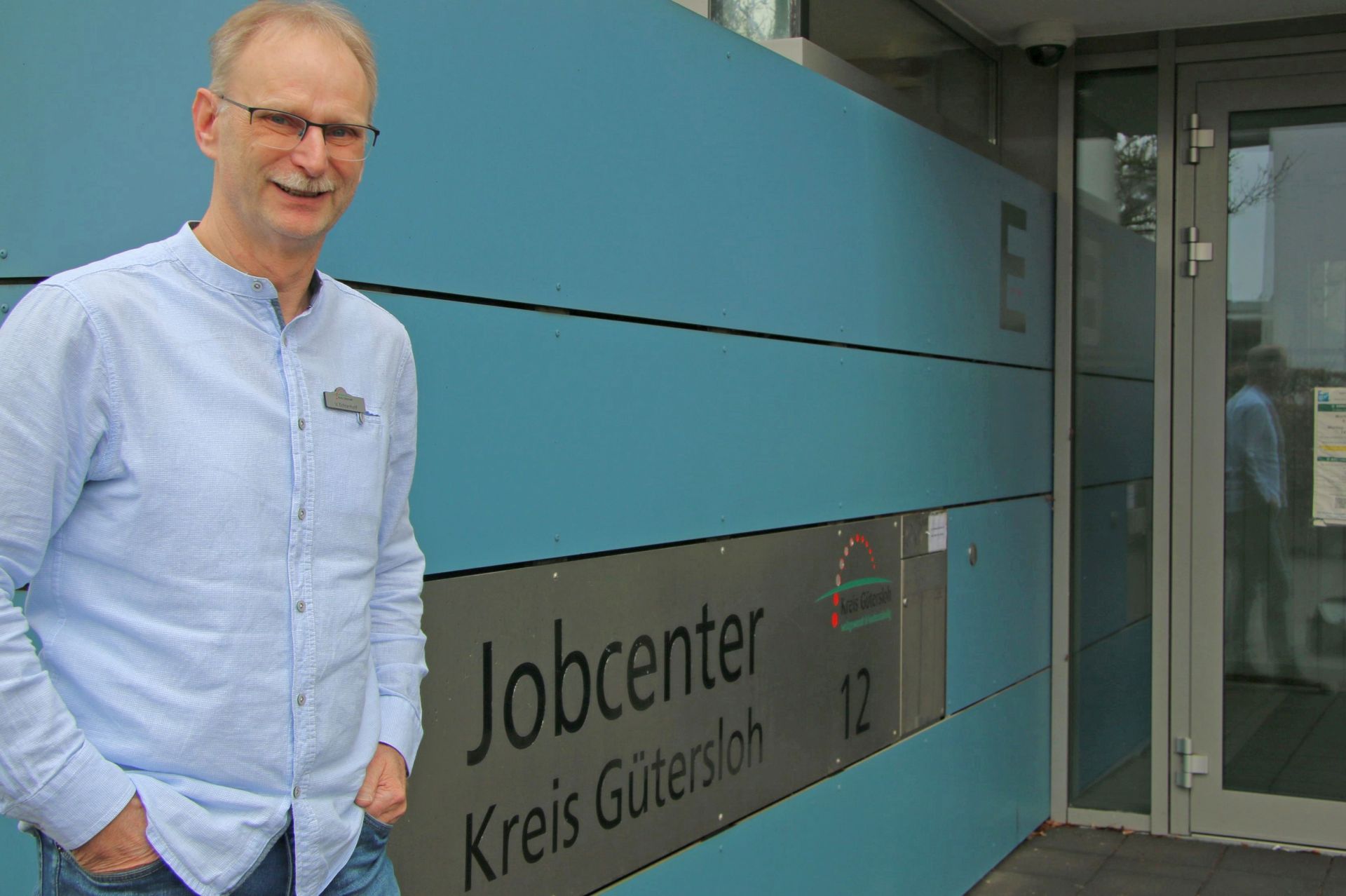 Ein Mann ist links im Bild. Er steht vor einer blauen Wand, die auf den Eingang des Jobcenters Kreis Gütersloh zuführt.