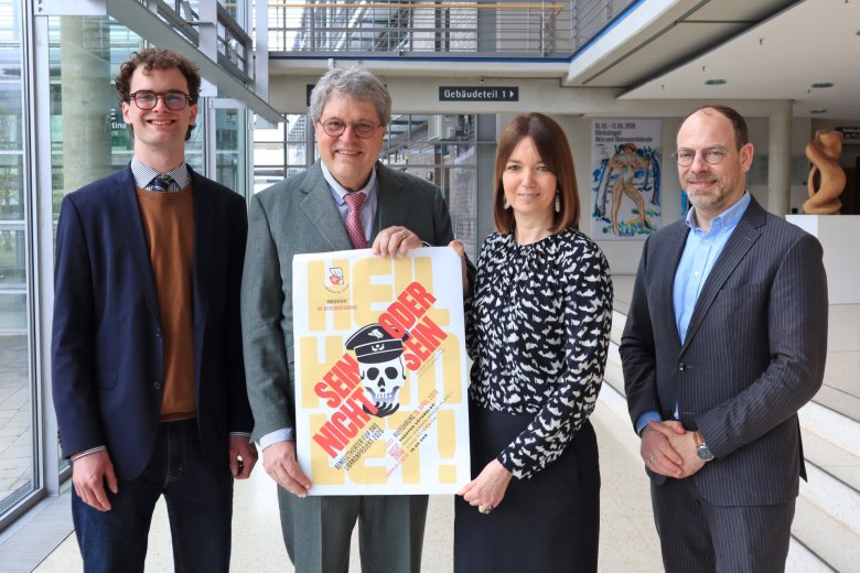 Vier Personen stehen nebeneinander. Die beiden Personen in der Mitte halten ein Plakat in den Händen. 