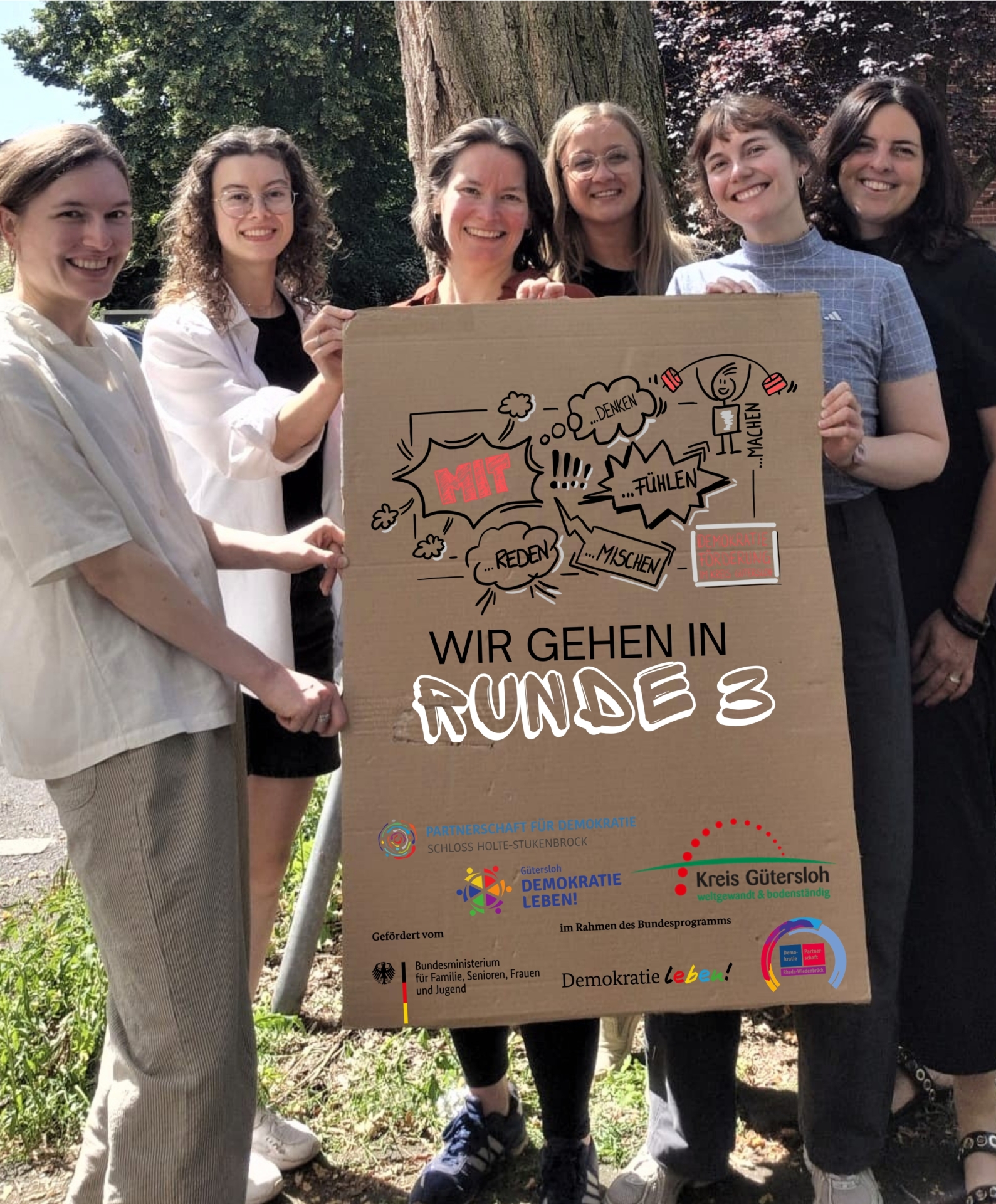 Wir gehen in Runde 3. - 1 Sechs Personen stehen nebeneinander und halten ein Plakat zu dem Projekt in den Händen.