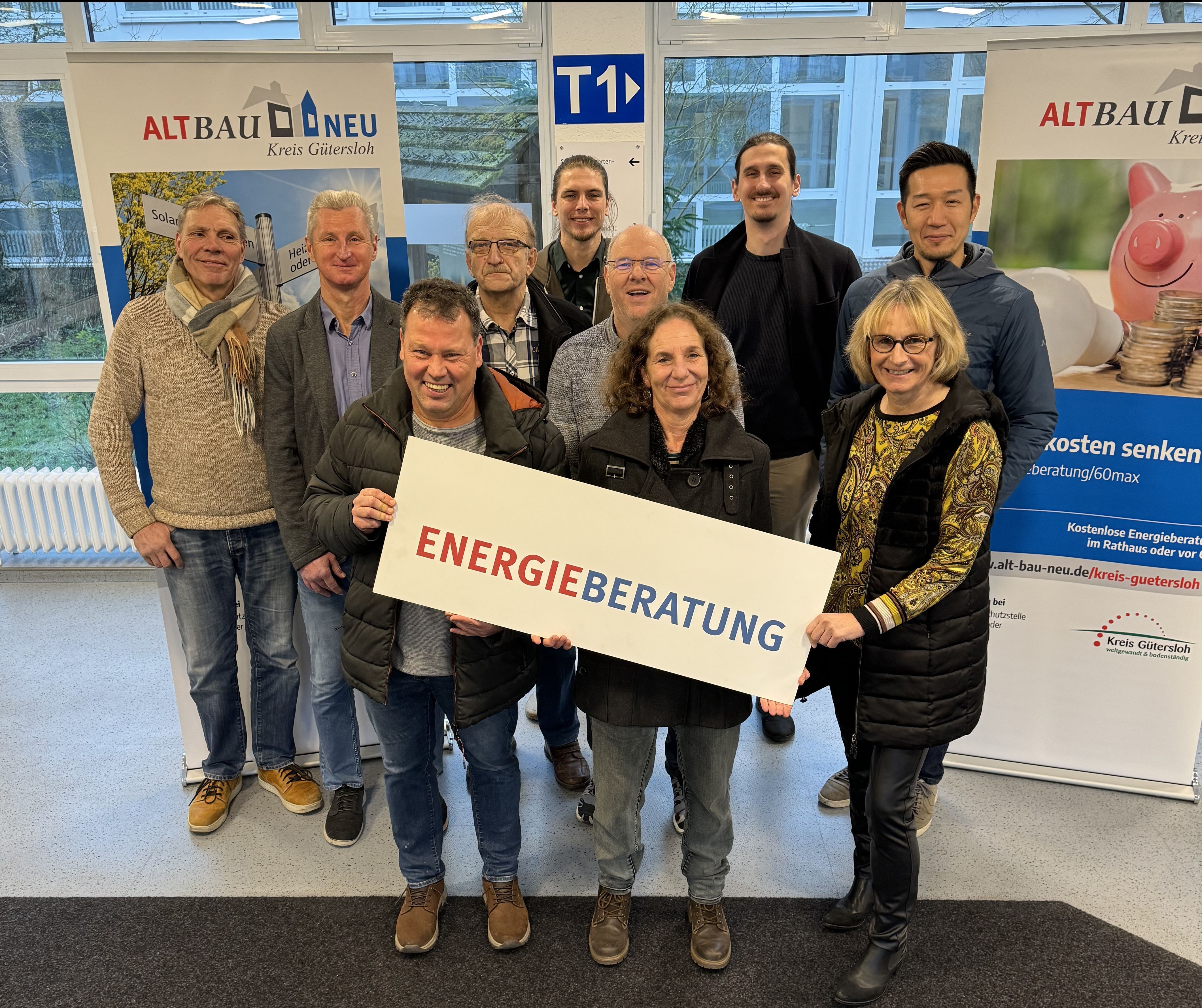 Personen stehen nebeneinander und blicken in die Kamera. Sie halten ein Banner mit der Aufschrift 'Energieberatung' in den Händen. 