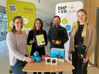 4 Personen stehen nebeneinander. Zwei halten jeweils einen kleinen Roboter in der Hand. Eine Person ein Ipad und eine weitere ein Podcast-Mikrofon.