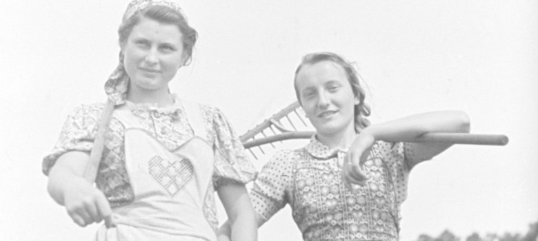 Zwei Frauen stehen nebeneinander auf einem Feld und halten Arbeitsgeräte in den Händen.