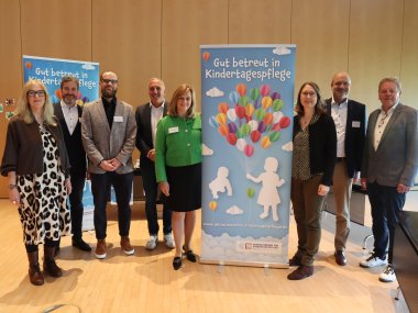Acht Personen stehen in einem Raum, in der Mitte ist ein blaues Banner über Kindertagespflege.