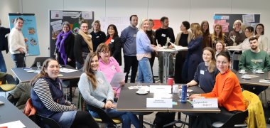 Raum gefüllt mit Menschen, die entweder stehen oder sitzen. Manche schreiben auch etwas an eine Flipchart. 