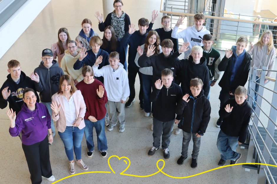 Ein Foto mit 23 Personen aus der Vogelperspektive im Foyer des Kreis Gütersloh. Der Großteil der Menschen sind Jugendliche. Im unteren Bereich ist eine Herzgrafik zu sehen.
