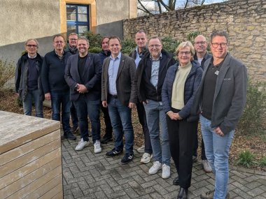 Eine Gruppe von Menschen steht vor einer Mauer und blickt in die Kamera.