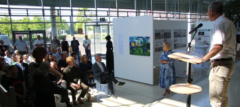 Ausstellung der Design- und Kunstschule Valmiera im Kreishaus Gütersloh Landrat Adenauer am Rednerpult im Foyer des Kreishauses, vor ohm die Besucherinnen und besucher.