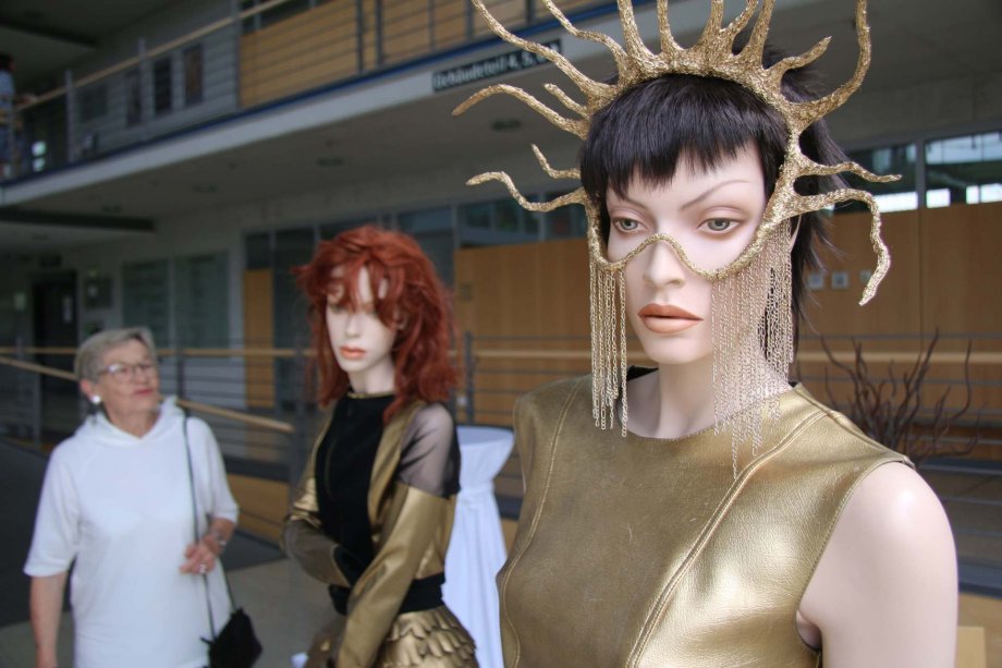 Zwei Schaufensterpuppen mit Entwürfen der schülerinnen und Schüler der Design- und Kunstschule Valmiera und eine Bescuherin der Ausstellungseröffnung.