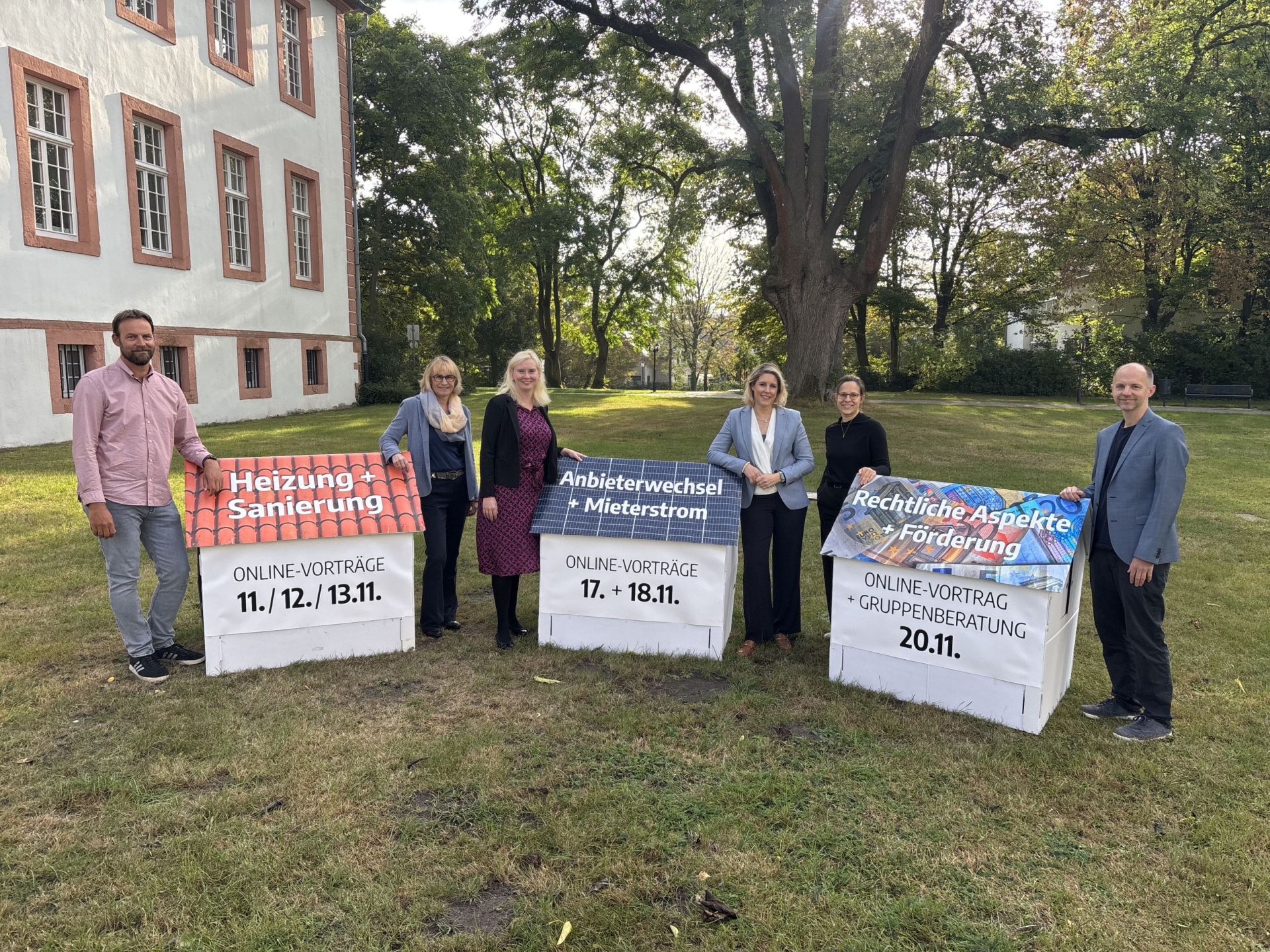 Personen stehen im Grünen, neben Papphäusern auf denen Daten und Titel der Vorträge stehen. 