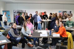 Raum gefüllt mit Menschen, die entweder stehen oder sitzen. Manche schreiben auch etwas an eine Flipchart. 