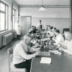 Schwarz-weiß Foto: Männer mit Mikroskopen und Lupen sitzen an einem langen Tisch. 
