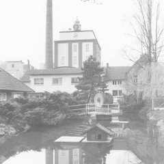 Schwarz-weiß Foto: Fabrikgebäude mit Schornstein und dem Firmenlogo "FMV", im Vordergrund ein Teich. 