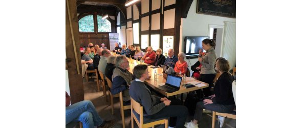 Im Mai fand eine Geschichtswerkstatt im Heimathaus Isselhorst statt. Mehrere Personen sitzen an einem Tisch, die Gütersloher Stadtarchivarin Julia Kuklik steht und referiert.