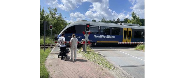 Das Bild zeigt einen Zug der Nordwestbahn mit Sitz in Osnabrück am Bahnübergang Carl-Benz-Straße in Steinhagen. Vor der geschlossenen Bahnschranke wartet eine Familie mit Kinderwagen.