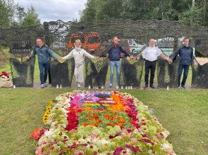 Mehrere Teilnehmer stehen in einem metallenen Monument mit Aussparungen in Menschenform und halten sich an den Händen. Im Vordergrund sind Blumengestecke.